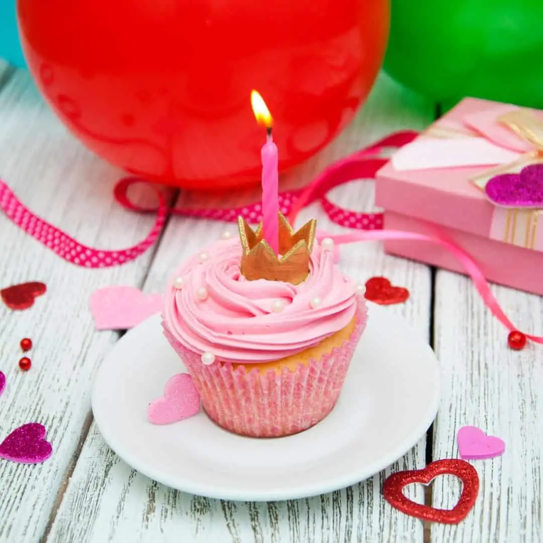 online virtual birthday party. image of cupcate on white plate. pink wrapper, pink frosting pink candle with small gold princess crown. red balloon and pink wrapped gift in background with heart shaped confetti scattered throughout.