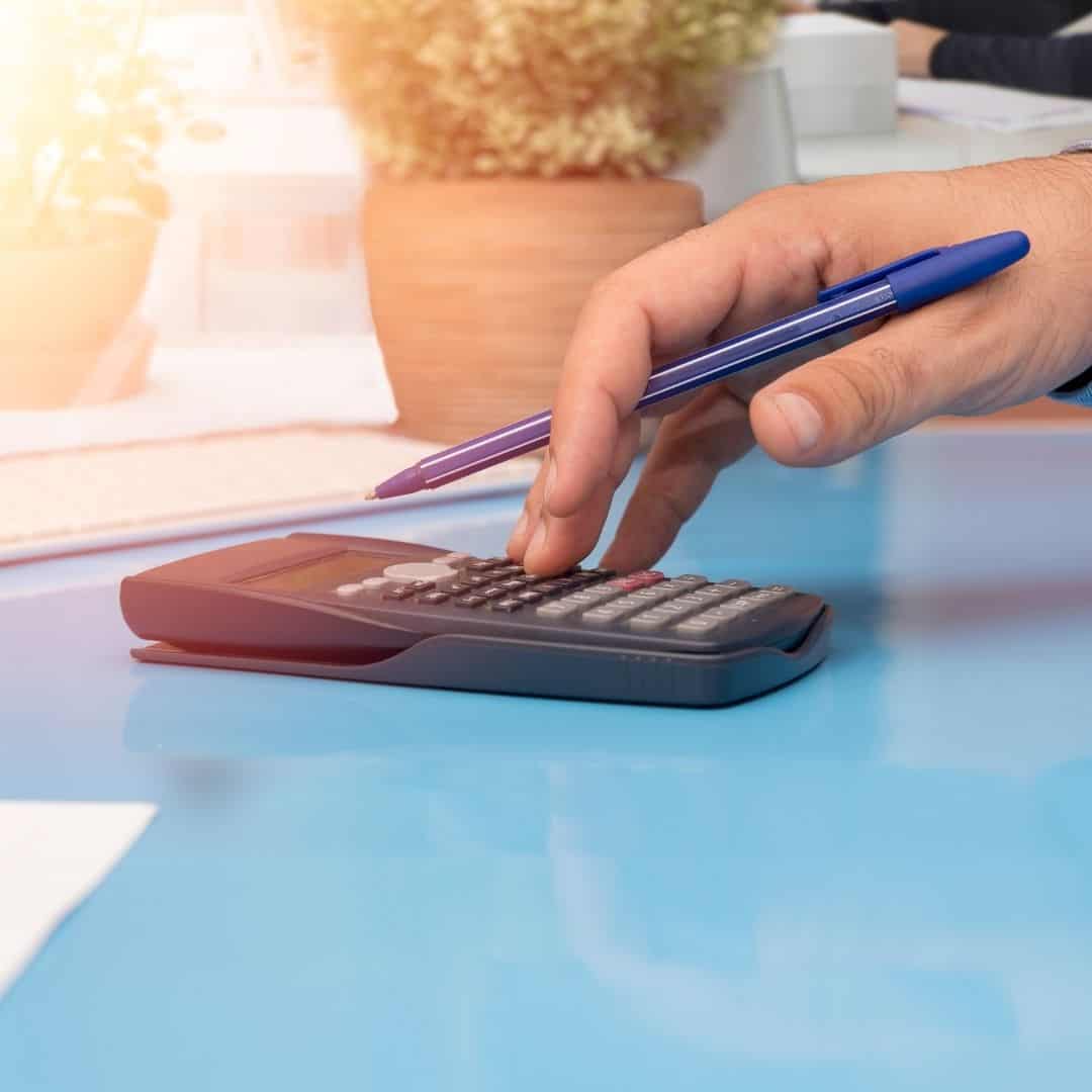 add up expenses, necessities; how to budget when unemployed. image of calculator on blue glass table top with hand calculating while holding pen.
