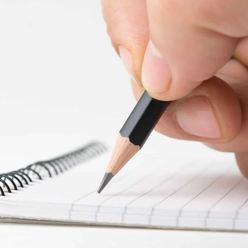 be informed is the best tip for how to survive a layoff financially. ask many questions, take notes. image of hand holding pencil depicting writing on lined paper in notebook