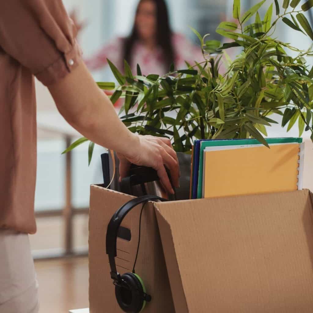 article is about how to survive a layoff financially, be professional not emotional when being laid off from work.  image depicts a woman leaving with a packed box and plant saying bye to femal co-worker