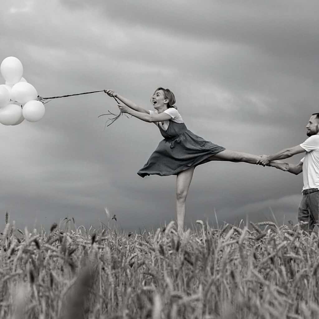 image: man holding woman trying to hold back a bundle of balloons. black and white